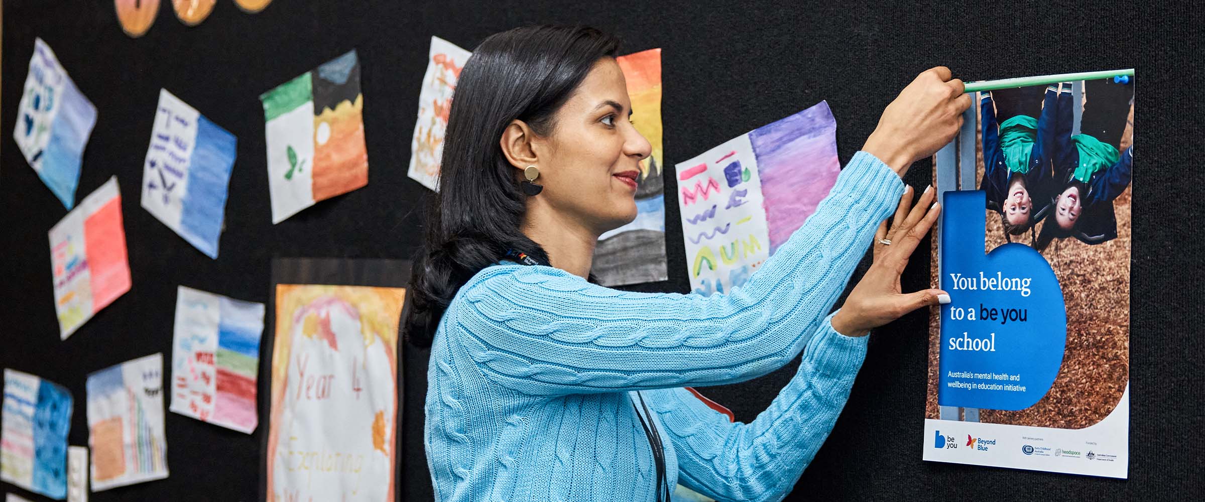 Teacher pinning a Get Started Be You poster on the staffroom noticeboard for colleagues.