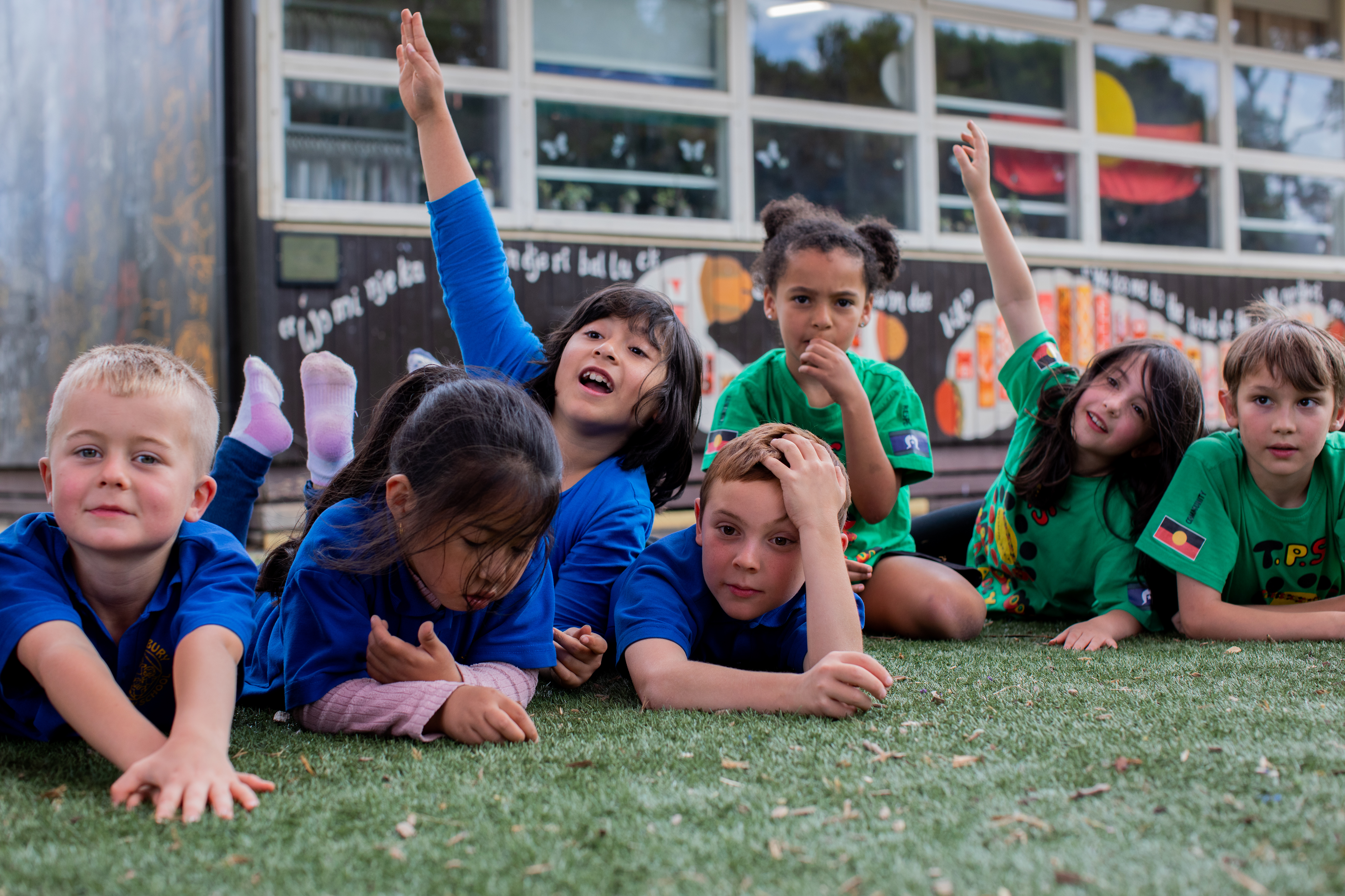 A group of seven primary school students laying and sitting on the grass in front of a classroom.