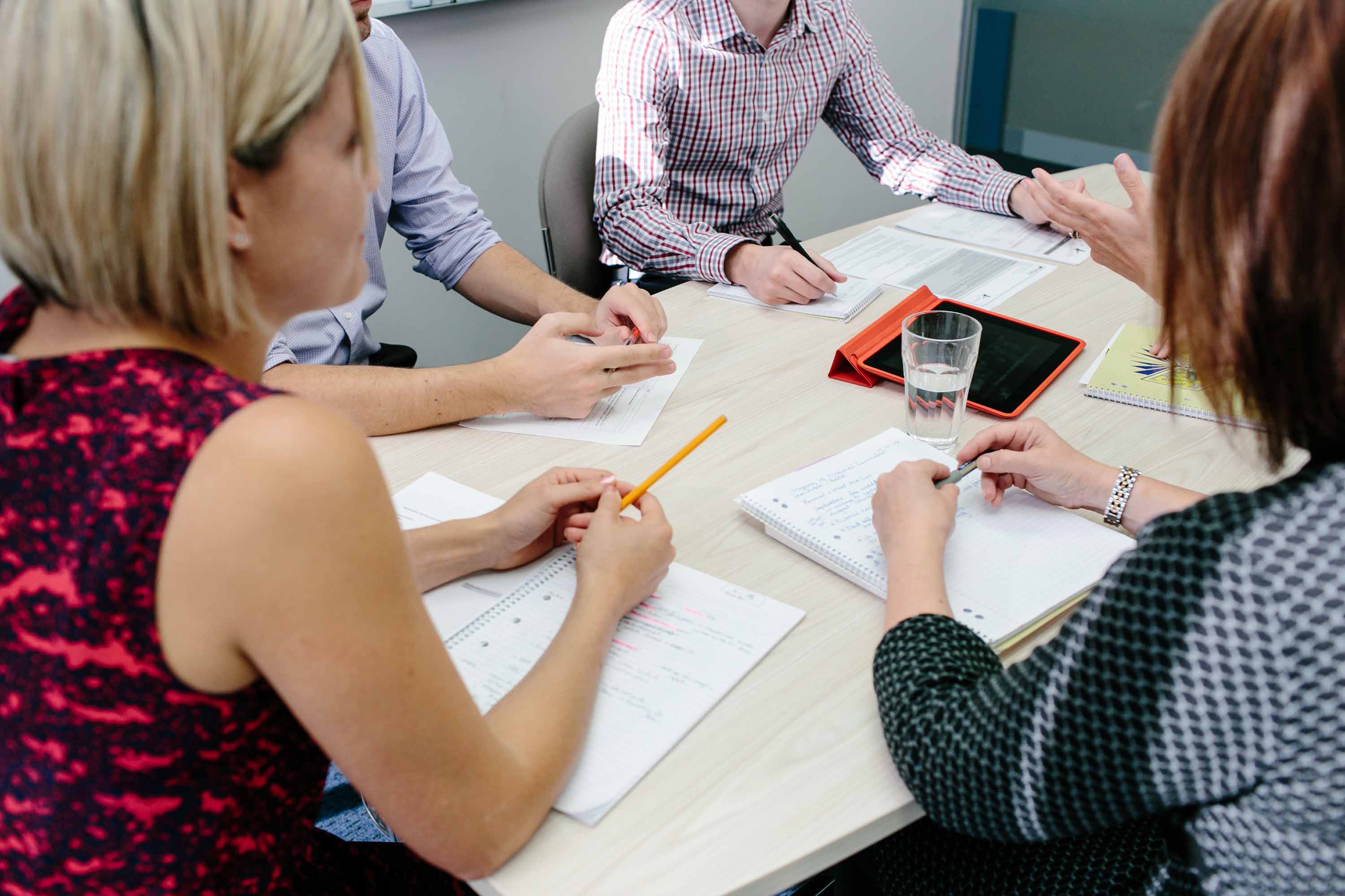 A group of four educators sitting around a table. There a notepads, glasses of water and a tablet on the table.