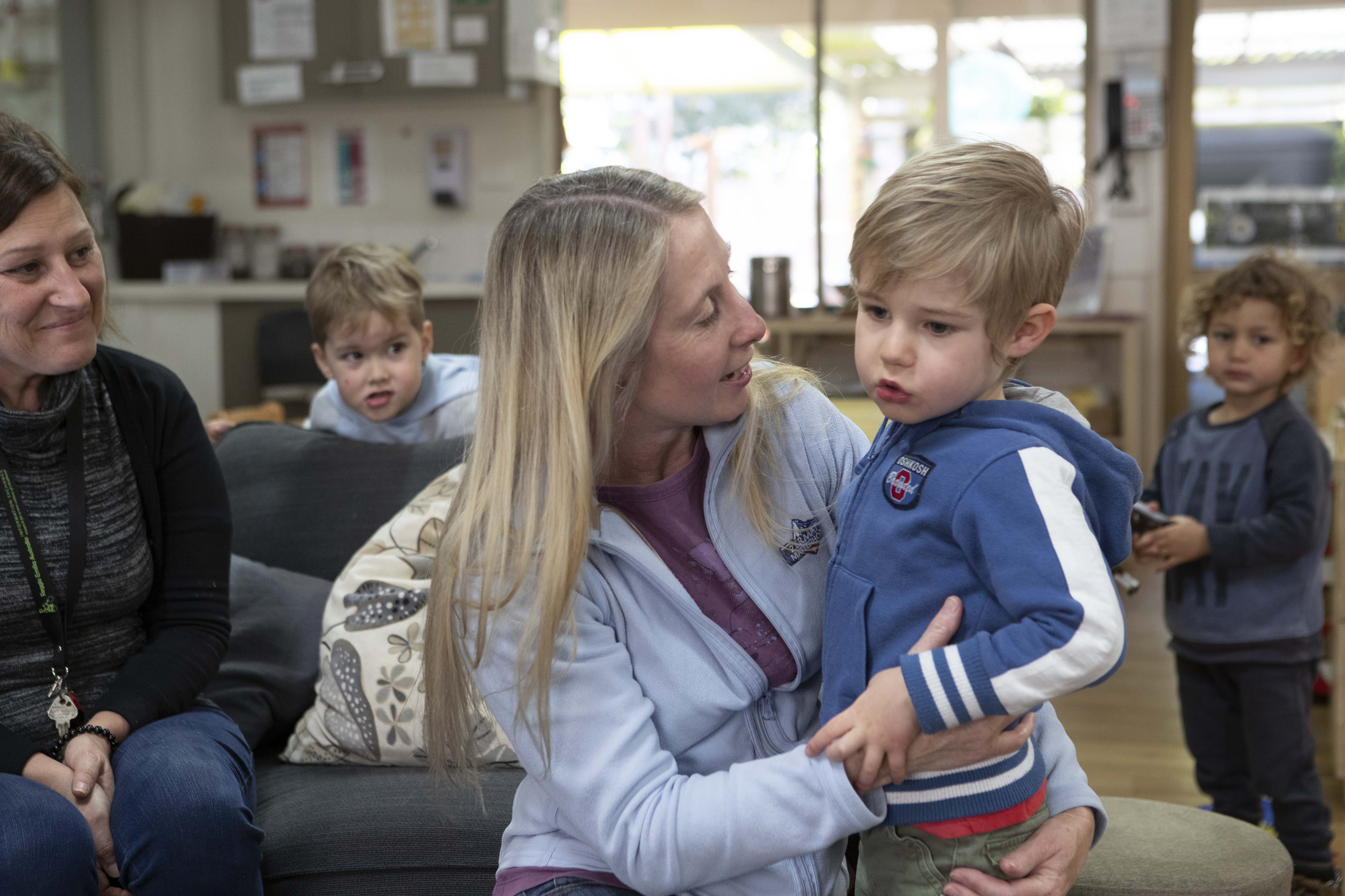 An adult is kneeling on the floor holding a young child. There is another adult and children in the background.