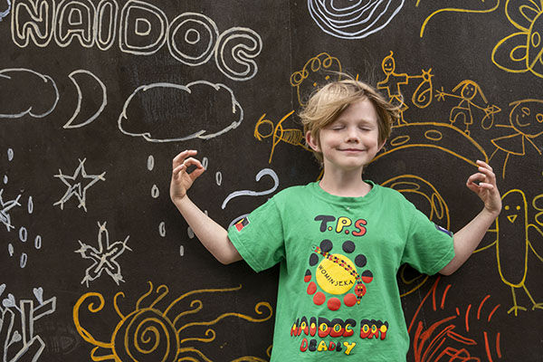 A young child in front of a chalk-board decorated for NAIDOC, with hands in meditation posture